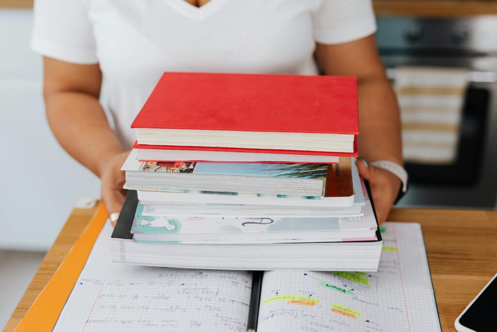 pexels-photo-5386433-5386433 Close-up of a person holding a stack of colorful books over an open notebook.